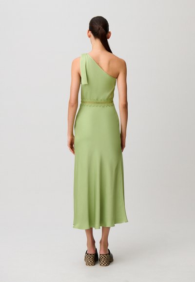 Donna con coda di cavallo scura che indossa un vestito midi verde chiaro con una spalla scoperta e scarpe con tacco a blocco decorate, vista di schiena.