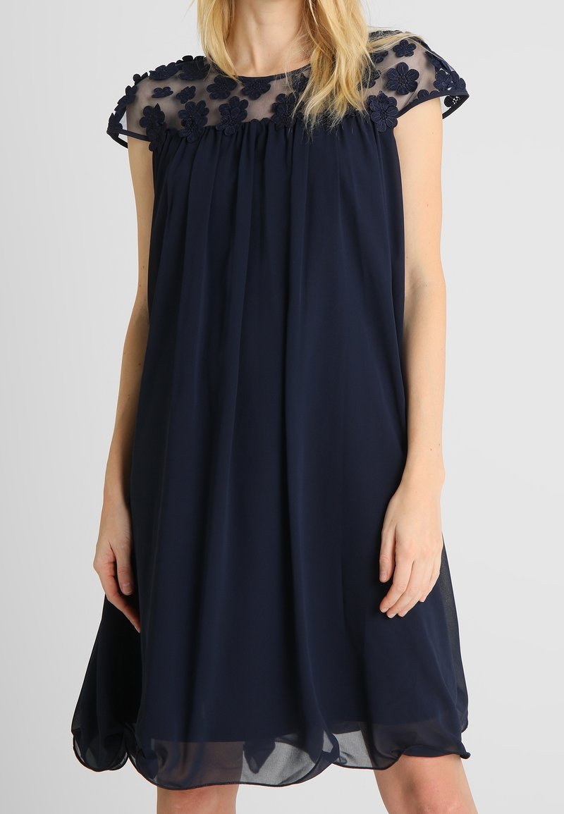 Femme portant une robe bleu marine sans manches avec la partie supérieure de la poitrine transparente et des détails d'appliqués floraux sur le décolleté.