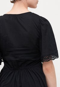 Schwarzes Kleid mit kurzen Puffärmeln, einem runden Ausschnitt und bestickten Punkten. Der Saum hat aufwendige Spitzenverzierungen. Weicher, strukturierten Stoff.