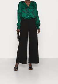 Grönmönstrad blus med V-hals och långa ärmar, kombinerad med höga svarta vida byxor och svarta öppna sandaler.