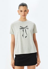 Kvinde med slicked-back hår iført hoops øreringe, en lysegrå t-shirt med et sort bånddesign og en sort nederdel foran en hvid baggrund.