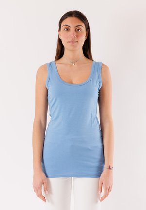 Donna con i capelli lunghi e scuri, che indossa un top azzurro chiaro senza maniche e pantaloni bianchi, in piedi contro uno sfondo bianco uniforme.