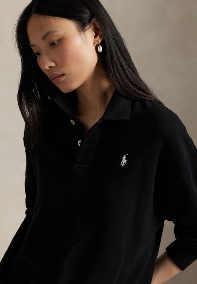 Young woman with long dark hair wearing gold hoop earrings and a black collared sweater with white logo on chest, looking down.