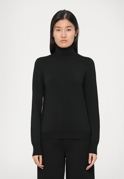 Woman standing facing forward wearing a black turtleneck sweater and black pants against a plain light background.