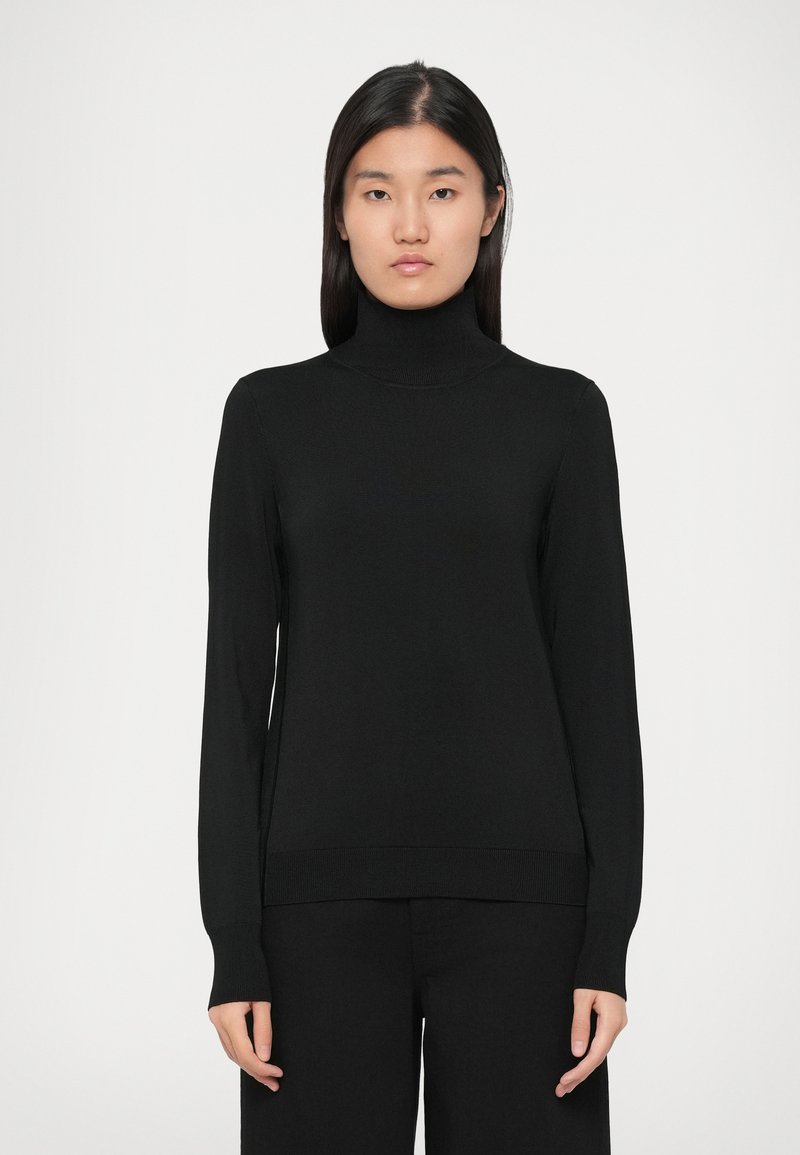 Kvinde står med ansigtet fremad iført en sort højhalset sweater og sorte bukser foran en ensartet lys baggrund.