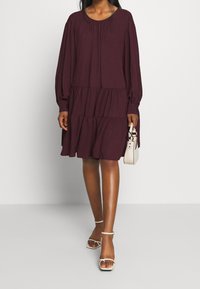 Vínová vícerozměrná šaty s dlouhými rukávy a kulatým výstřihem. Měkká tkanina s volným designem. Doplněno o bílé sandály na podpatku a malou kabelku.
