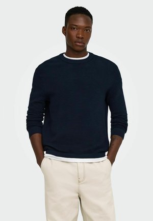 Homme aux cheveux courts et bouclés portant un pull côtelé bleu foncé sur une chemise blanche et un pantalon beige clair, debout les mains dans les poches.
