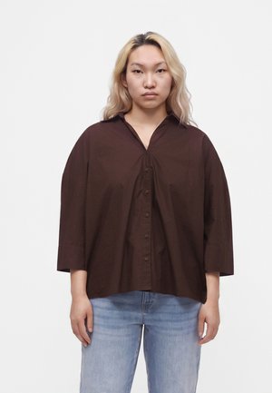 Ung person med skulderlangt blondt hår iført en løs, mørkebrun skjorte med knapper og lyseblå jeans, stående mod en hvid baggrund.