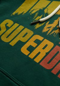 Zielona bluza z dużym żółtym i czerwonym napisem „SUPERDRY”, z grafiką góry i drzewa. Ma kremowy sznurek.
