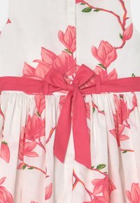 Vestido sin mangas en tela blanca con un estampado de flores rosas. Cuenta con una cinturilla rosa con un lazo atado en la parte delantera y una falda recogida.