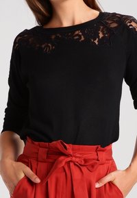 Pull noir avec détails en dentelle sur les épaules, col rond et manches trois-quarts. Associé à un pantalon rouge plissé taille haute.