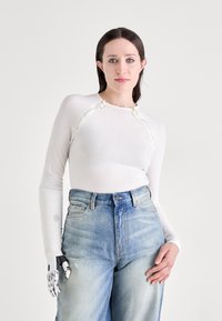 Weißes Longsleeve-Oberteil mit rundem Ausschnitt und dekorativen, kreisförmigen Akzenten. Kombiniert mit hochtaillierten, hellblauen Jeans.