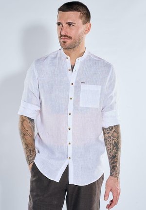 Mann mit Tattoos trägt hochgekrempeltes weißes Langarmhemd und dunkle Hose, schaut zur Seite vor schlichtem hellem Hintergrund.