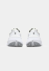 Chaussures de sport blanches avec une tige texturée, un talon renforcé et un col rembourré. La semelle crantée blanche et grise offre de la stabilité. Détails réfléchissants présents.