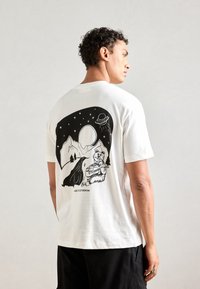 Witte katoenen t-shirt met een zwarte grafische illustratie van bergen, een beer en een UFO op de achterkant. Korte mouwen, relaxte pasvorm.