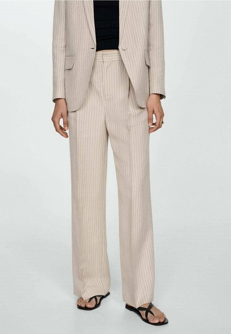 Pantalon beige à fines rayures, coupe ample, associé à un blazer assorti, un haut noir et des sandales noires ouvertes à fines lanières.