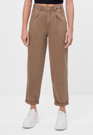 Osoba ubrana w brązowe, zwężane spodnie z wysokim stanem z podwiniętymi mankietami oraz białe sneakersy na platformie, stojąca na tle jednolitego, białego tła.