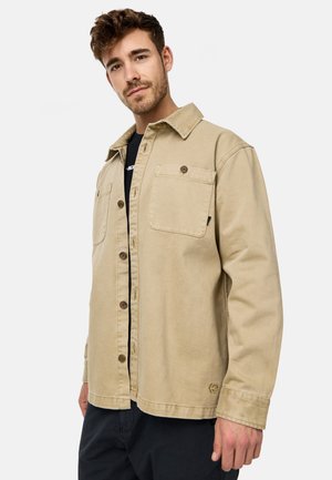 Beige cotton shirt jacket with a button-up front, two chest pockets, and a collar, featuring visible stitching and a logo detail on the hem.