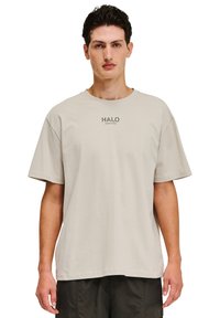 Lys beige bomulds t-shirt med en afslappet pasform. Har korte ærmer og en rund halsudskæring. Grafisk "HALO" tekst trykt i mørkt på brystet.