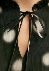 Zwarte geplooide blouse met een keyhole halslijn, voorzien van een ruffle-kraag en verstelbare touwtjes. Gekleed met grote witte stippen die in de stof vervagen.