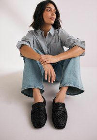 Zapatos negros de lona tipo slip-on con un diseño texturizado y dos acentos negros lisos. Combinados con jeans de pierna ancha de color azul claro y una camisa a rayas.