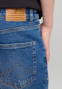 Sininen farkkuhousut, joissa on erityinen takataskujen muotoilu, oranssit tikkaukset ja vaaleanruskea nahkatee, jossa on kohokuvioitu teksti ja lippumalli.