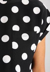 Svart tygblus med stora vita prickar, med en liten framficka och korta ärmar. Slät struktur och modern design.