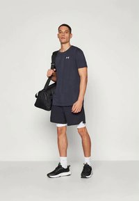 Vīrietis melnā sporta tērpā un baltās kompresijas šortos tur melnu sporta somu pār plecu, stāvot uz neizteiksmīga fona.