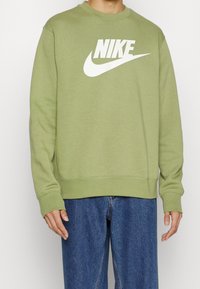 Ljus olivgrön sweatshirt med rund hals, prydd med en stor vit Nike-logotyp. Materialet verkar mjukt och designen är enkel.