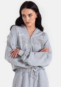 Szara krótka bluza rozpinana na zamek z przednim wzorem z kryształków, szerokimi rękawami i kieszenią typu kangur. Składa się w zestawie z dopasowanymi szarymi spodniami dresowymi.