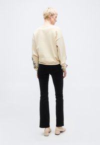 Άτομο με κοντά ξανθά μαλλιά που φοράει μπεζ φούτερ με μαύρα σχέδια στα μανίκια, μαύρο παντελόνι και μπεζ μποτάκια με τακούνι, γυρισμένο πλάτη.