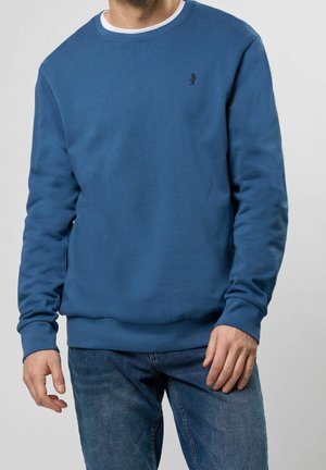 Felpa blu in morbido tessuto, con collo rotondo, polsini e orlo a costine, e un piccolo logo nero sul lato sinistro del petto. Indossata con jeans blu.