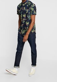 Kortærmet, marineblå skjorte med et grønligt og gult blomstermønster. Båret med mørke jeans og lysegule sneakers med hvide såler.