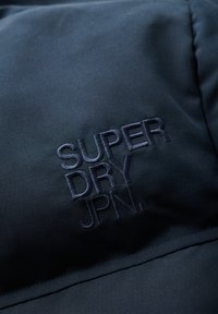 Tessuto nero con sottile trama, caratterizzato da ricamo "SUPER DRY JPN." in filo scuro. Vista ravvicinata dei dettagli del logo.