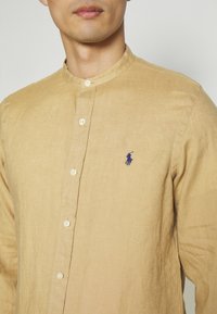 Mężczyzna ubrany w beżową koszulę na guziki z kołnierzykiem typu band i małym ciemnoniebieskim haftowanym logo polo z zawodnikiem na lewej piersi.