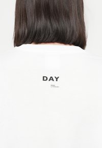 White cotton t-shirt with a round neckline, featuring a printed label "DAY" and brand name on the back. Simple, casual design.