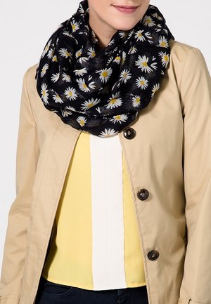 Femme portant une écharpe noire à motifs de marguerites blanches et jaunes, un manteau beige à boutons, et un chemisier jaune avec une bande blanche au centre.