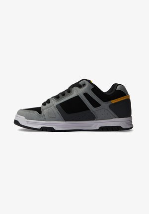 Zwarte en grijze sportieve schoen met een gestructureerde bovenkant, perforaties en een witte buitenzool met gele accenten op de hiel.