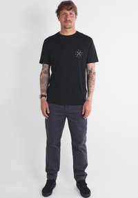 T-shirt en coton noir à manches courtes avec un logo circulaire sur la poitrine gauche. Associé à un pantalon gris et des baskets noires.