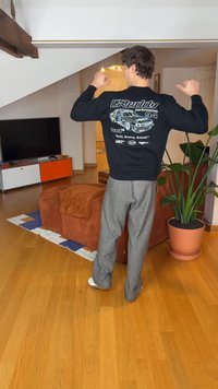 Čierna košeľa s dlhým rukávom s grafikou auta a textom na chrbte. Sivé textúrované nohavice. Vnútorné prostredie s hnedou pohovkou a rastlinami.