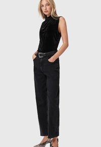 Jonge vrouw met blond haar, gekleed in een mouwloze zwarte fluwelen top, hoge zwarte jeans, een zwarte riem en metallic hakken, staand met haar handen in haar zakken.