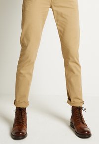 Botas de tobillo de cuero marrón con cordones y un acabado liso, combinadas con chinos beige arremangados en los puños, mostrando un estilo casual.