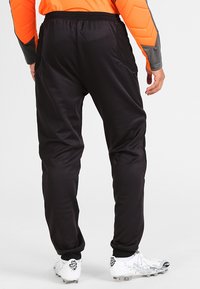 Schwarze Sporthosen mit glatter Textur, ausgestattet mit Seitentaschen und einem elastischen Bund, kombiniert mit weißen Fußballschuhen.