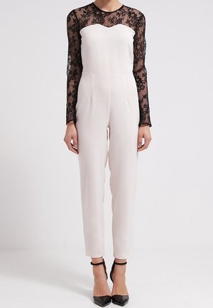 Mono blanco con un corsé ajustado y mangas largas de encaje negro transparente. Presenta un escote en forma de corazón y piernas de pantalón ceñidas, combinado con tacones negros.