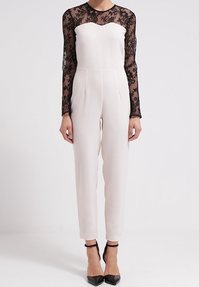 White jumpsuit with a fitted bodice and sheer black lace long sleeves. Features a sweetheart neckline and slim pant legs, paired with black heels.