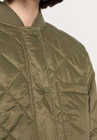Chaqueta acolchada verde oliva con cuello acanalado, cierre de botones y un bolsillo en el pecho, con una textura suave y un ligero brillo.