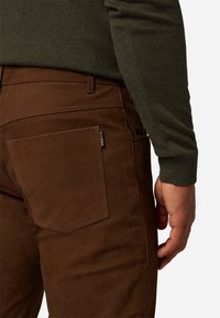 Bruine broek met een gladde textuur, voorzien van een detail in de achterzak en een label. De stof lijkt duurzaam en goed passend.