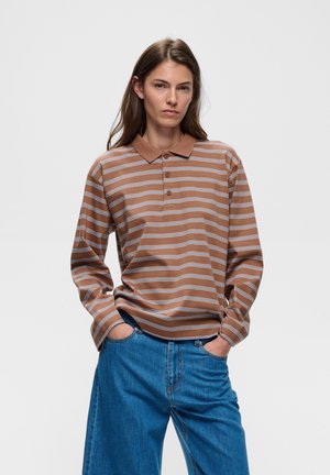 Ung person med langt hår iført en brun og hvid stribet langærmet poloshirt og blå jeans med hænderne i lommerne.