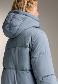Blondýna v světle modré zateplené zimní bundě s velkou kapucí, zobrazená zezadu a z boku.