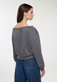 Szary, oversize'owy sweatshirt z szerokim dekoltem, luźnym krojem i ściągaczem u dołu. Połączony z niebieskimi dżinsami o wysokim stanie. Miękka faktura.